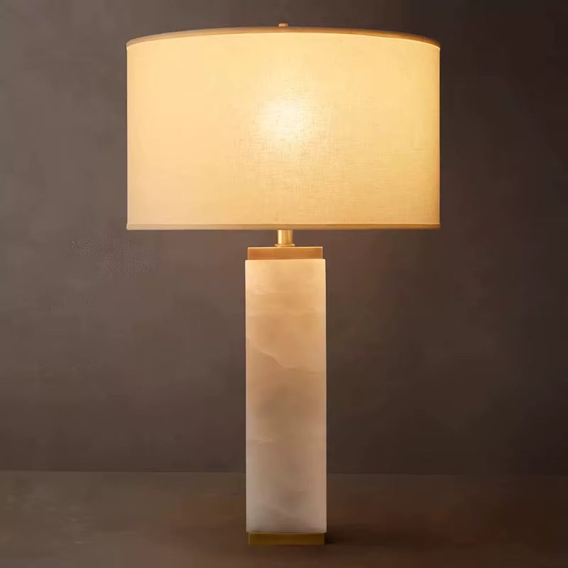 Serra white quartz column table lamp lit, with brass cap and ivory linen drum shade